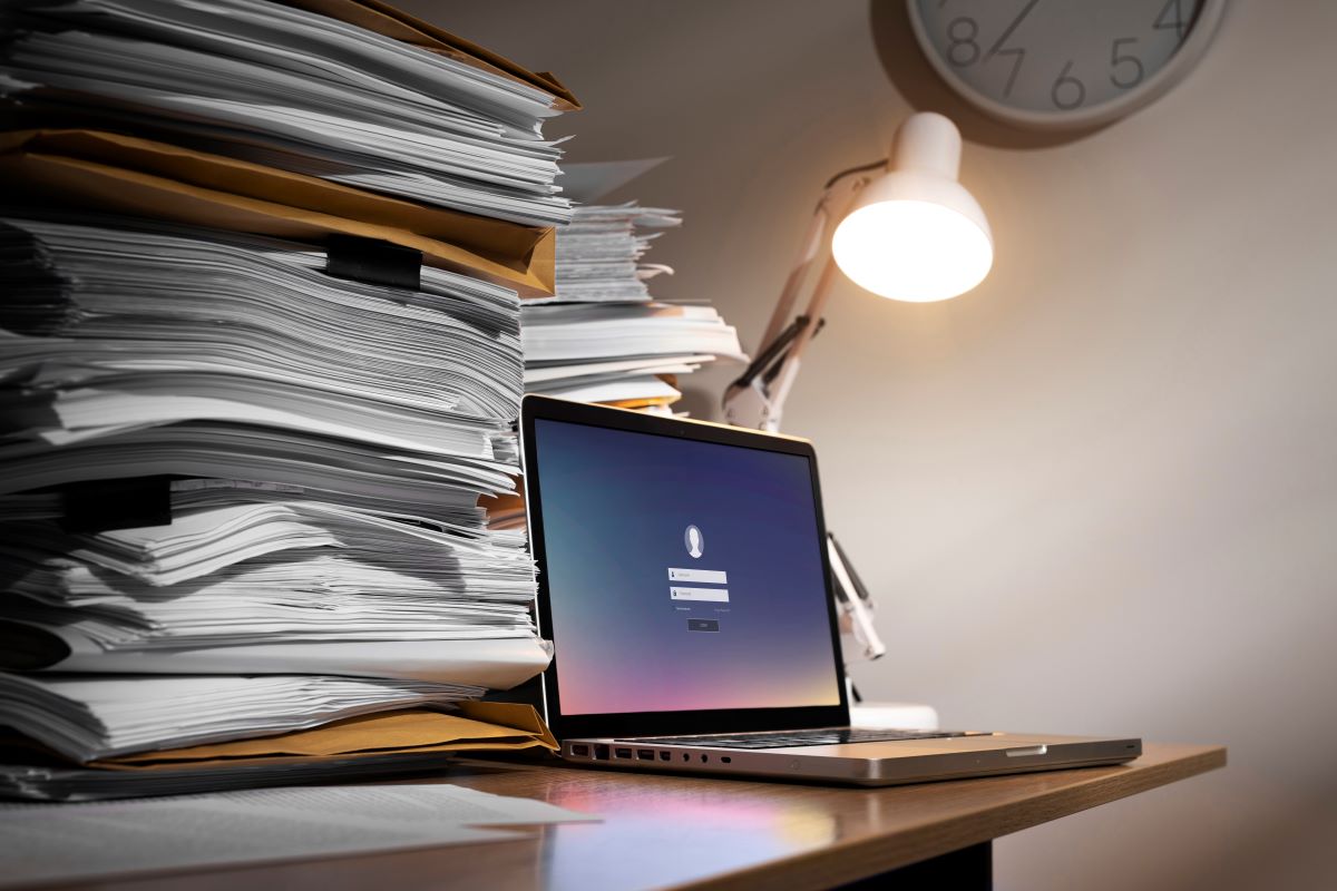 Zdjęcie przedstawia stos dokumentów i laptop.